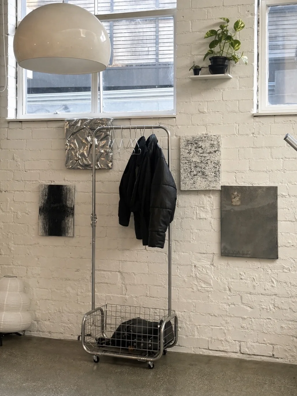 White-brick wall corner — chrome garment rack, small abstract artworks, a pendant lamp and a shelf plant