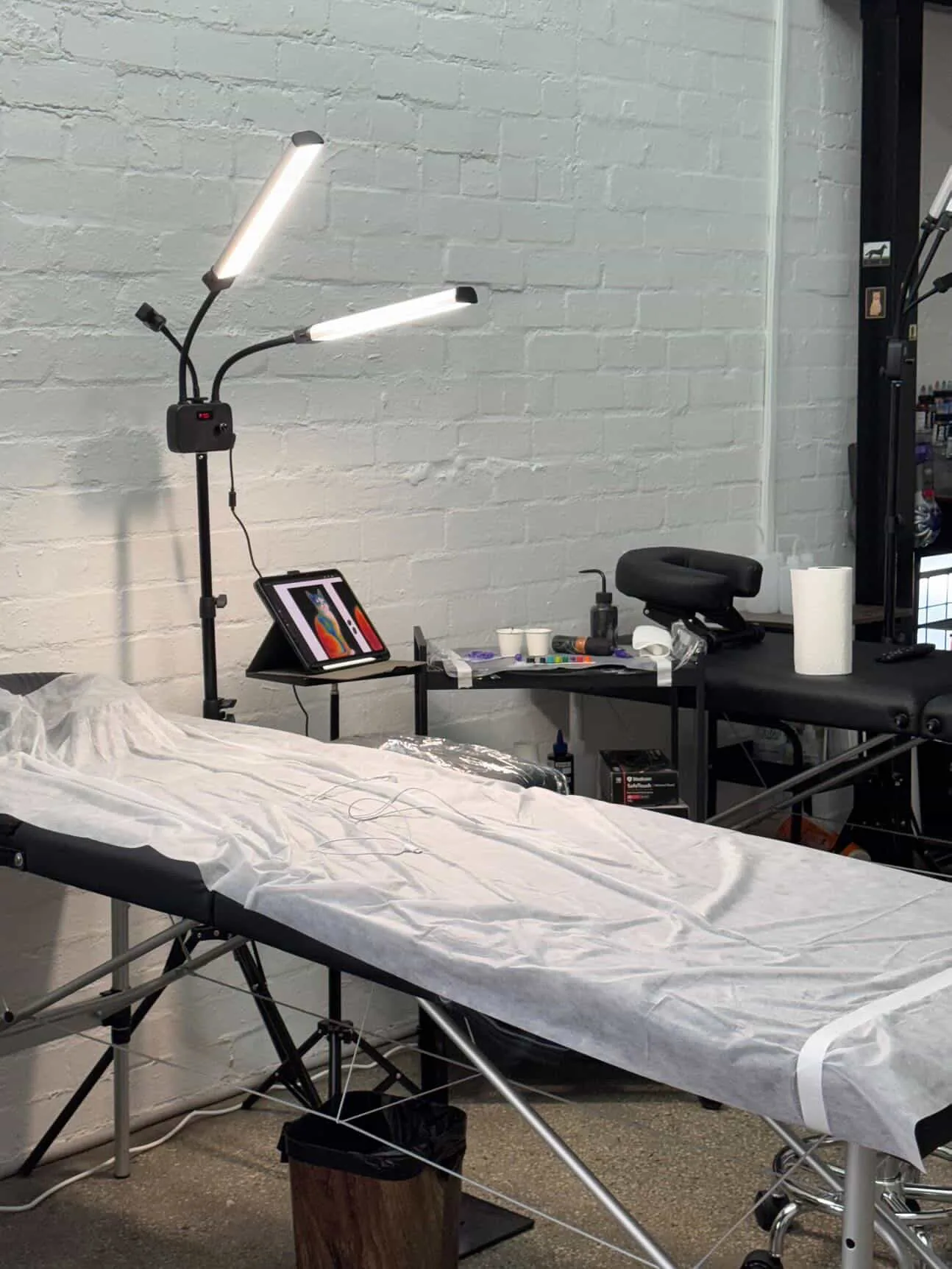 A set-up tattoo workstation — bed with barrier sheet, twin adjustable LED lamps, ink caps and tools on a black cart
