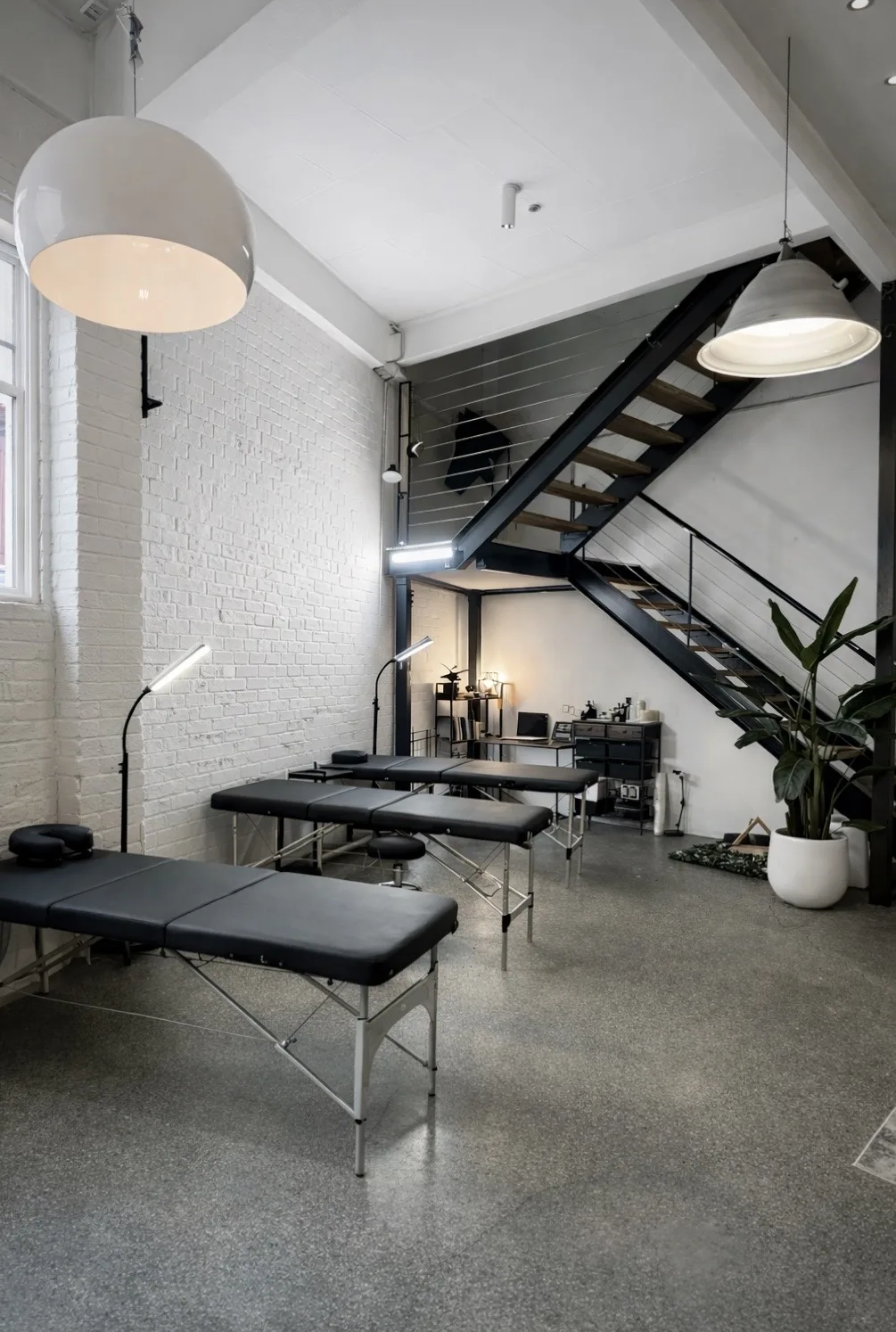 Wide loft interior — black steel staircase to mezzanine, white-brick walls, black tattoo tables and pendant lamps