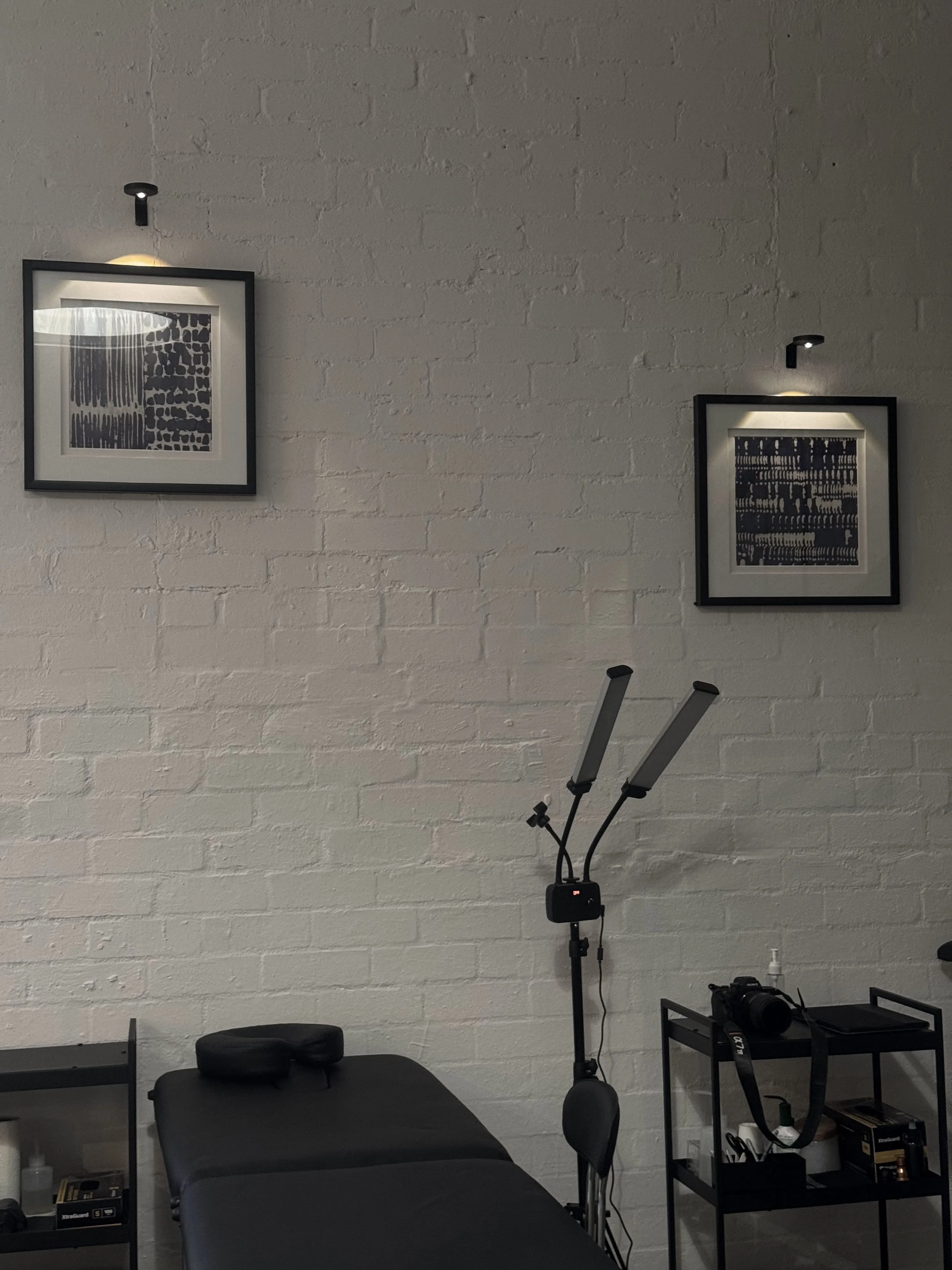 Interior corner — framed monochrome abstract artworks above a tattoo bed, twin LED lamp and white-brick wall