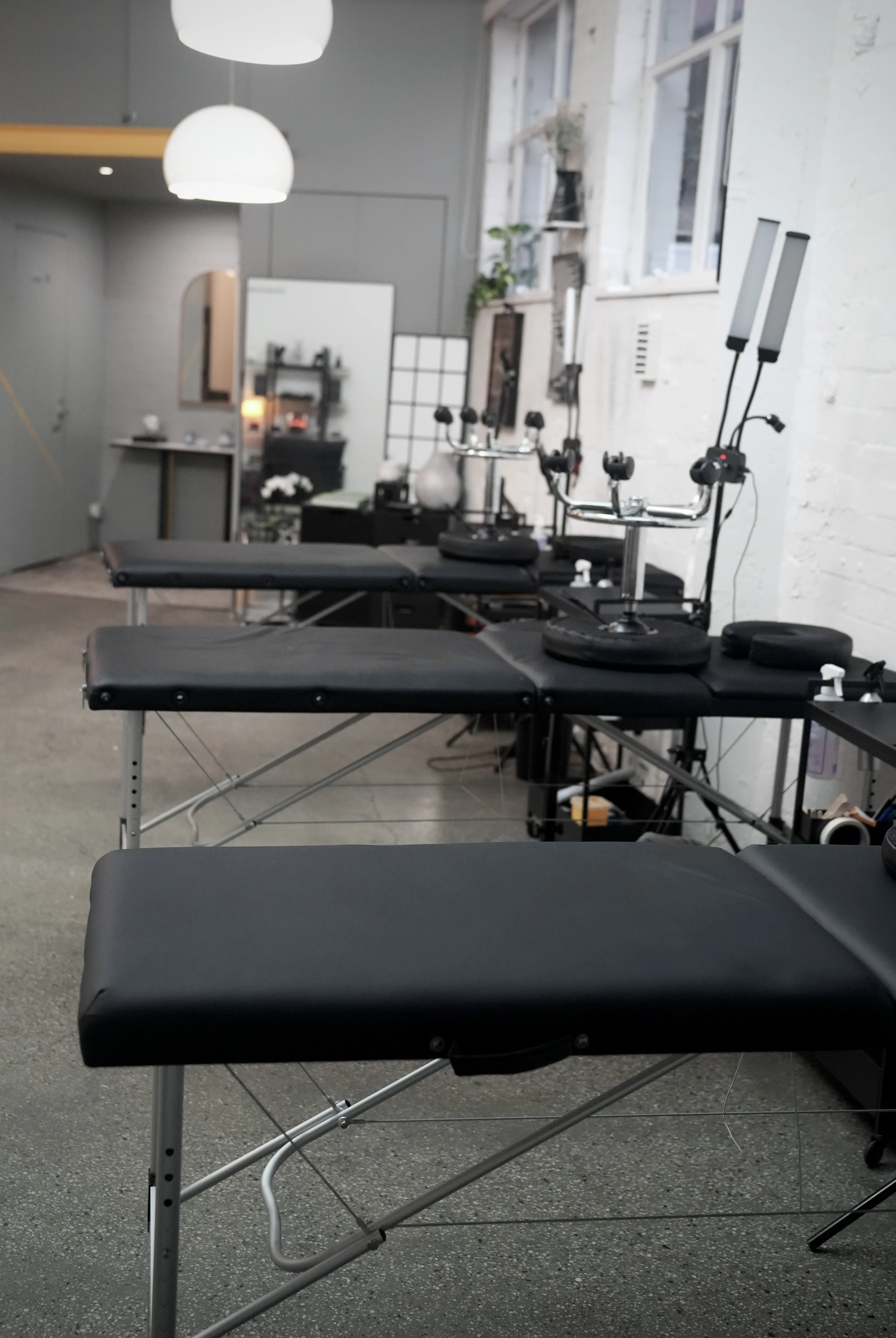 Row of black tattoo beds receding down the length of the studio — chrome arm-rest stands, pendant lamps and polished concrete floor